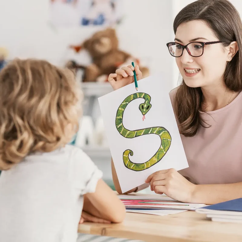 A female speech and language therapist holding a picture of a snake shaped like an "S" working with a young girl