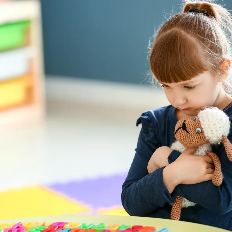 A little girl wth selective mutism sitting alone in a classroom hugging a stuffed toy