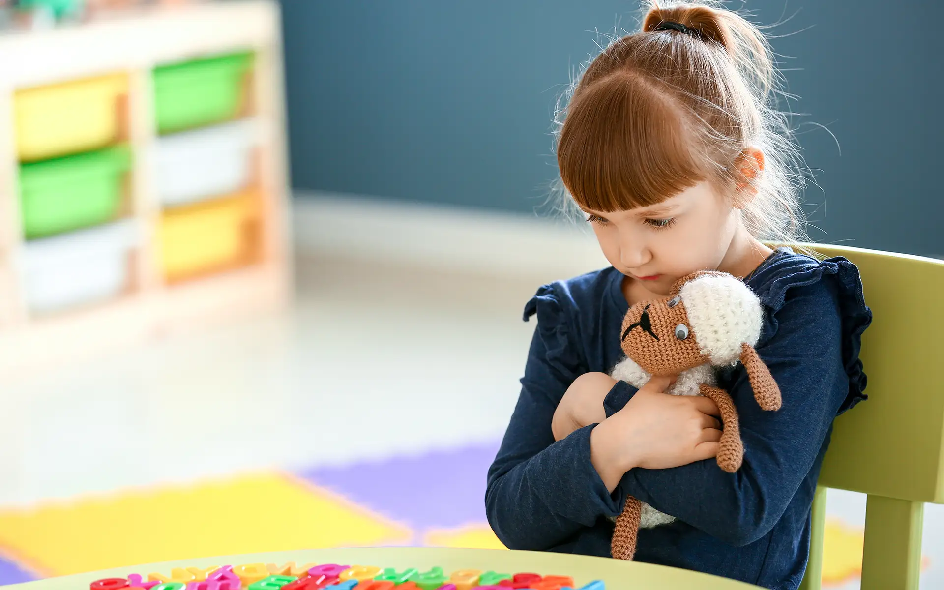 A little girl wth selective mutism sitting alone in a classroom hugging a stuffed toy