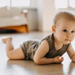 Baby on a wooden floor in a brown onesie, lying on its belly while looking up