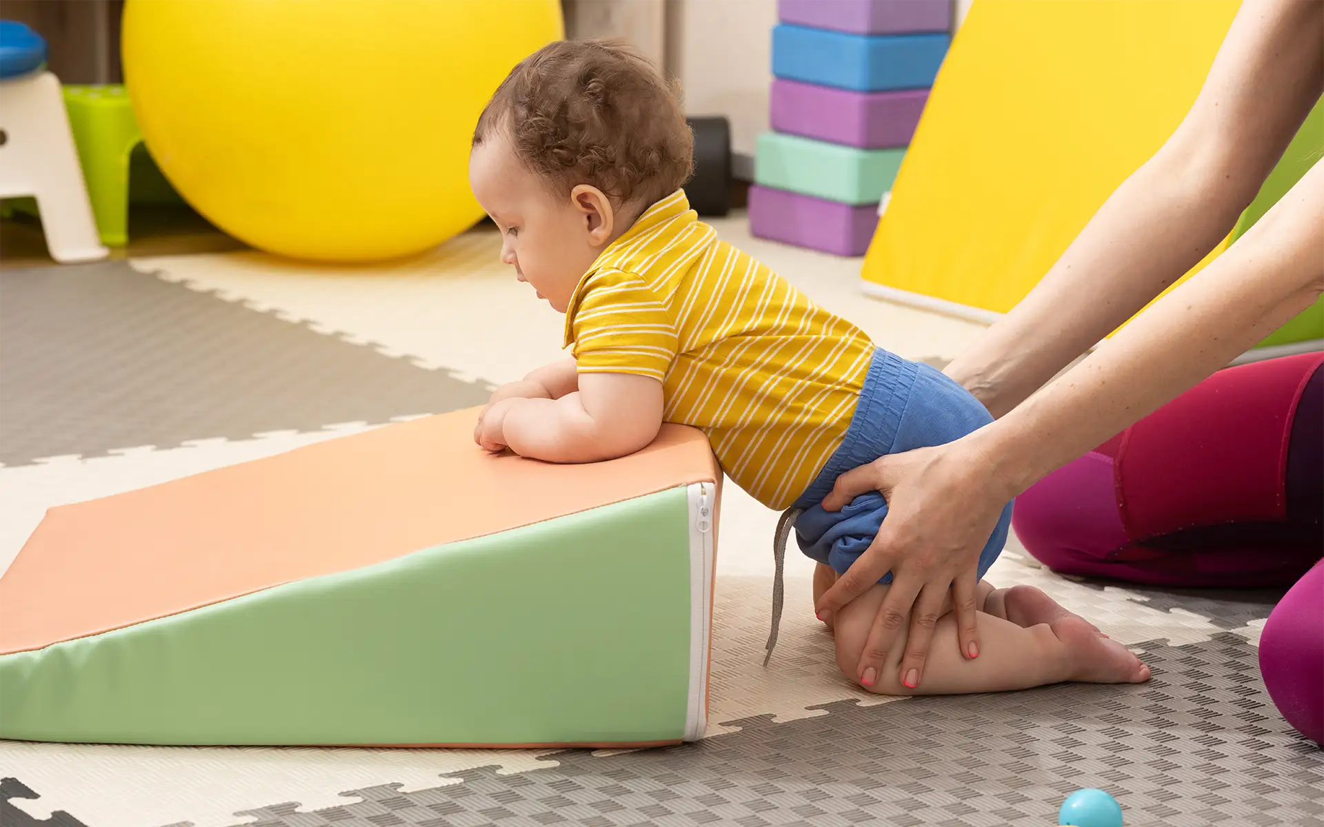 A pediatric physical therapist working with a toddler boy who is leaning over a therapy block