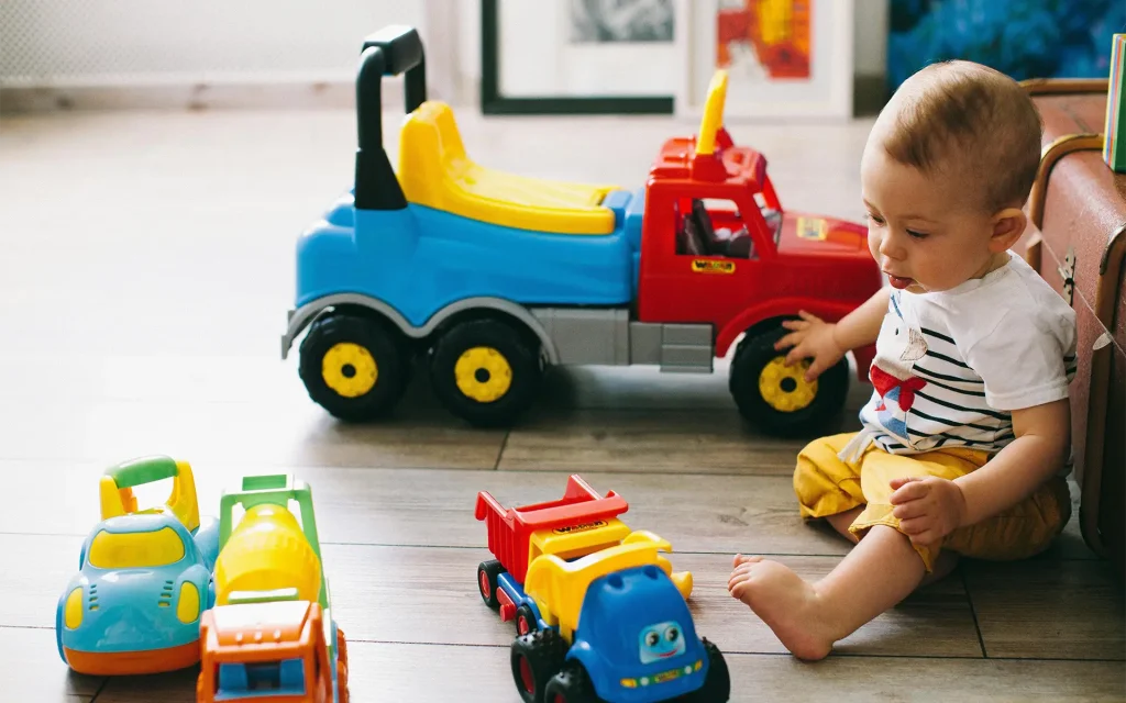 A baby boy is sitting in a safe place on the floor, looking at his toy trucks