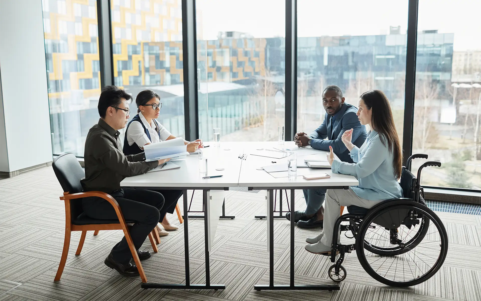 A diverse business team sits around a table in a conference room with windows, discussing a project with a focus on a young woman in a wheelchair