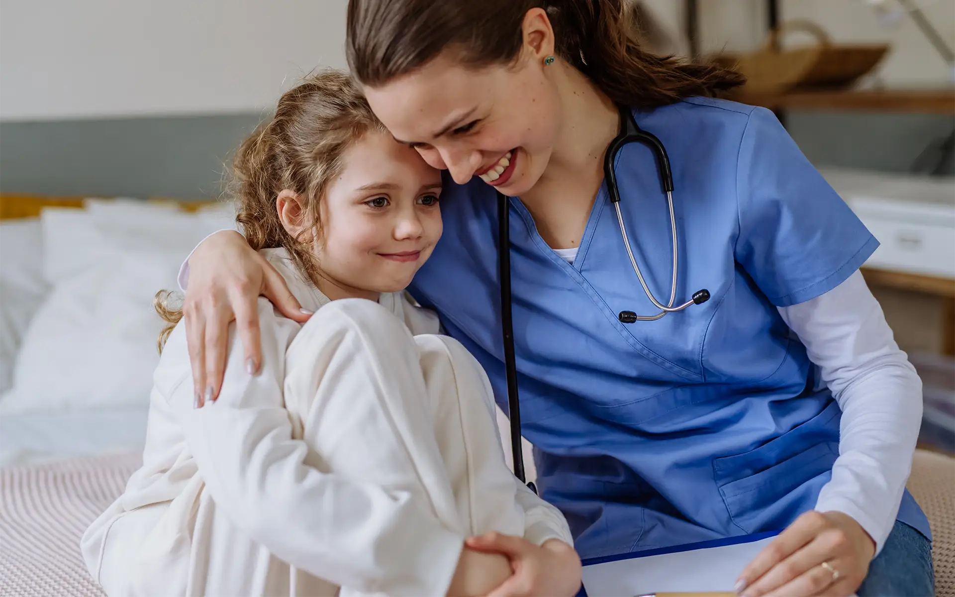 A female nurse hugging a smiling girl sitting in bed at home