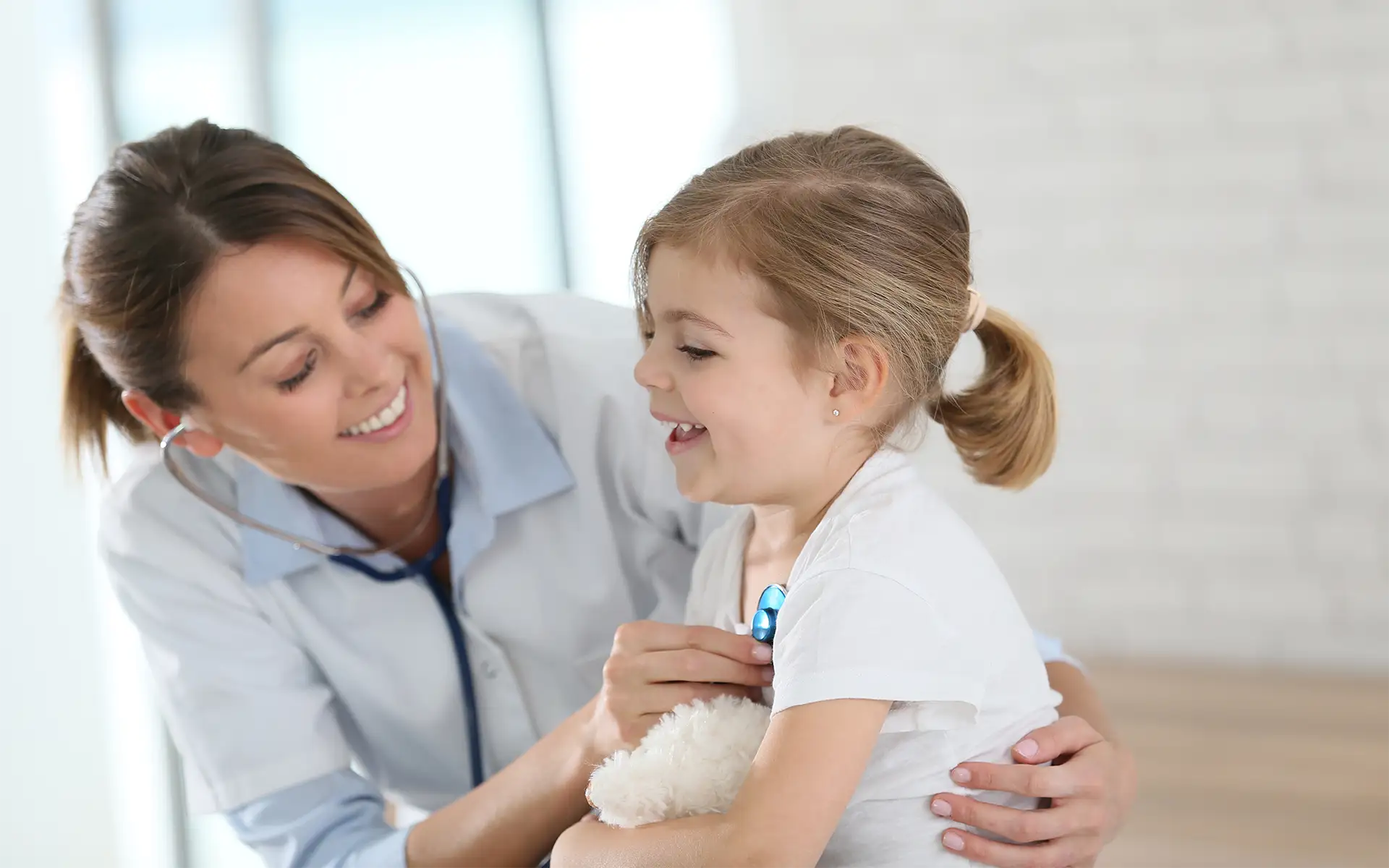 A female pediatric nurse smiles while examining a young girl patient using a stethoscope