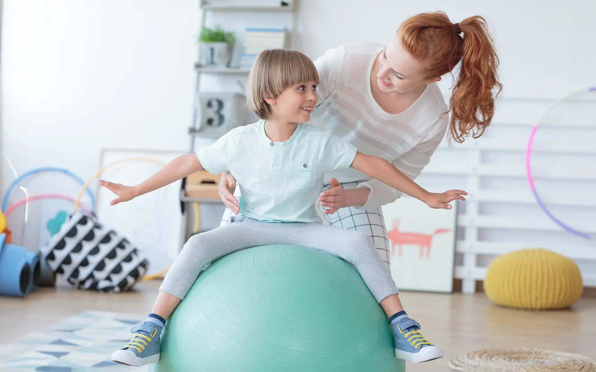 A female occupational therapist works with a young boy patient, helping him balance on a large green exercise ball
