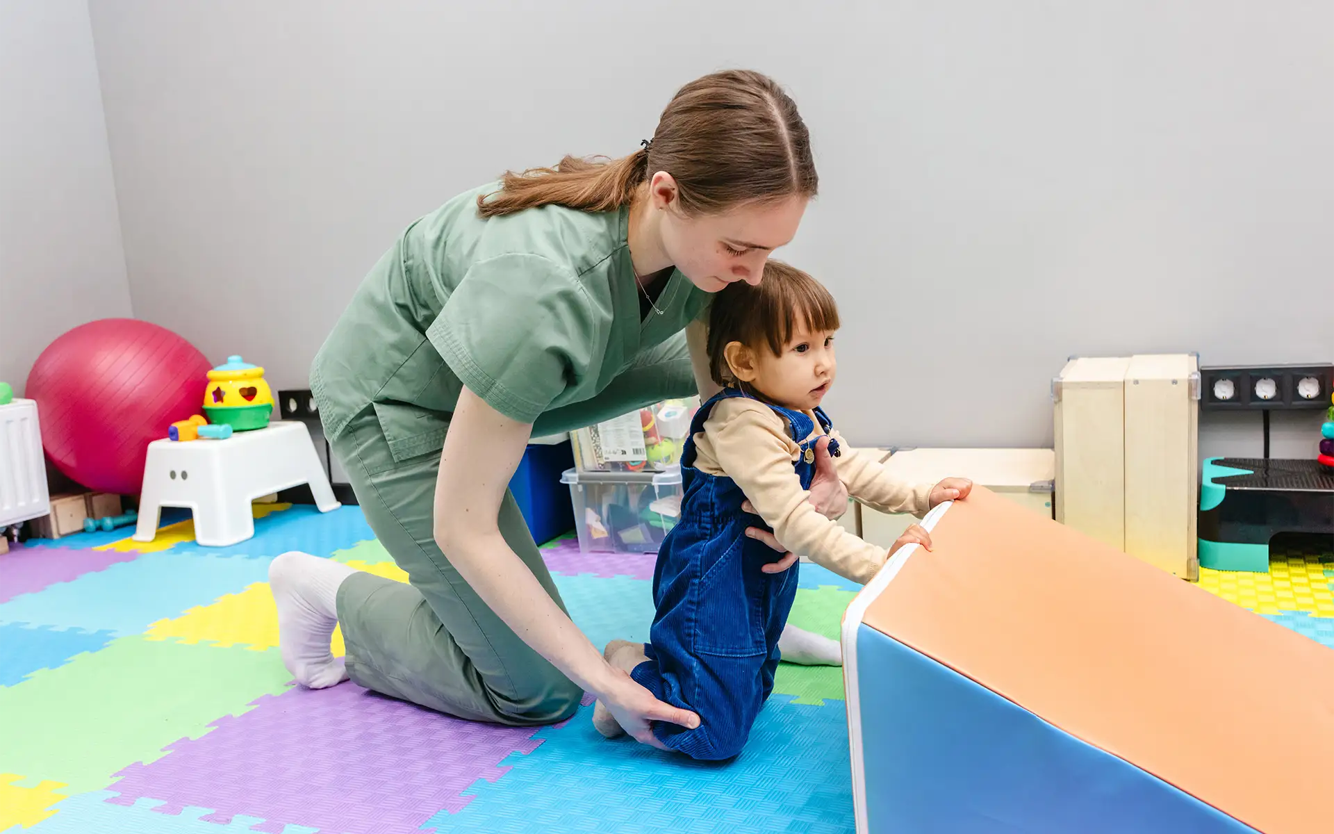 A pediatric physical therapist in green scrubs helps a female toddler with leg movement
