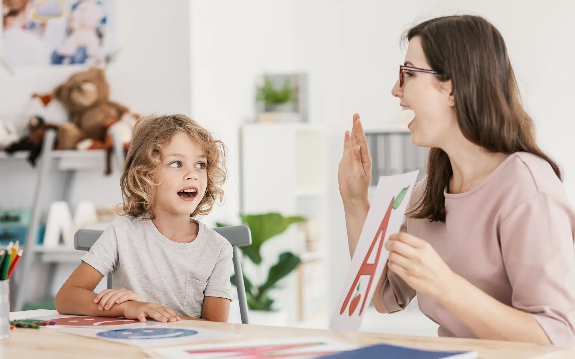 A pediatric speech language pathologist sits at a table with a young boy, holding up a letter "A" while working on pronunciation
