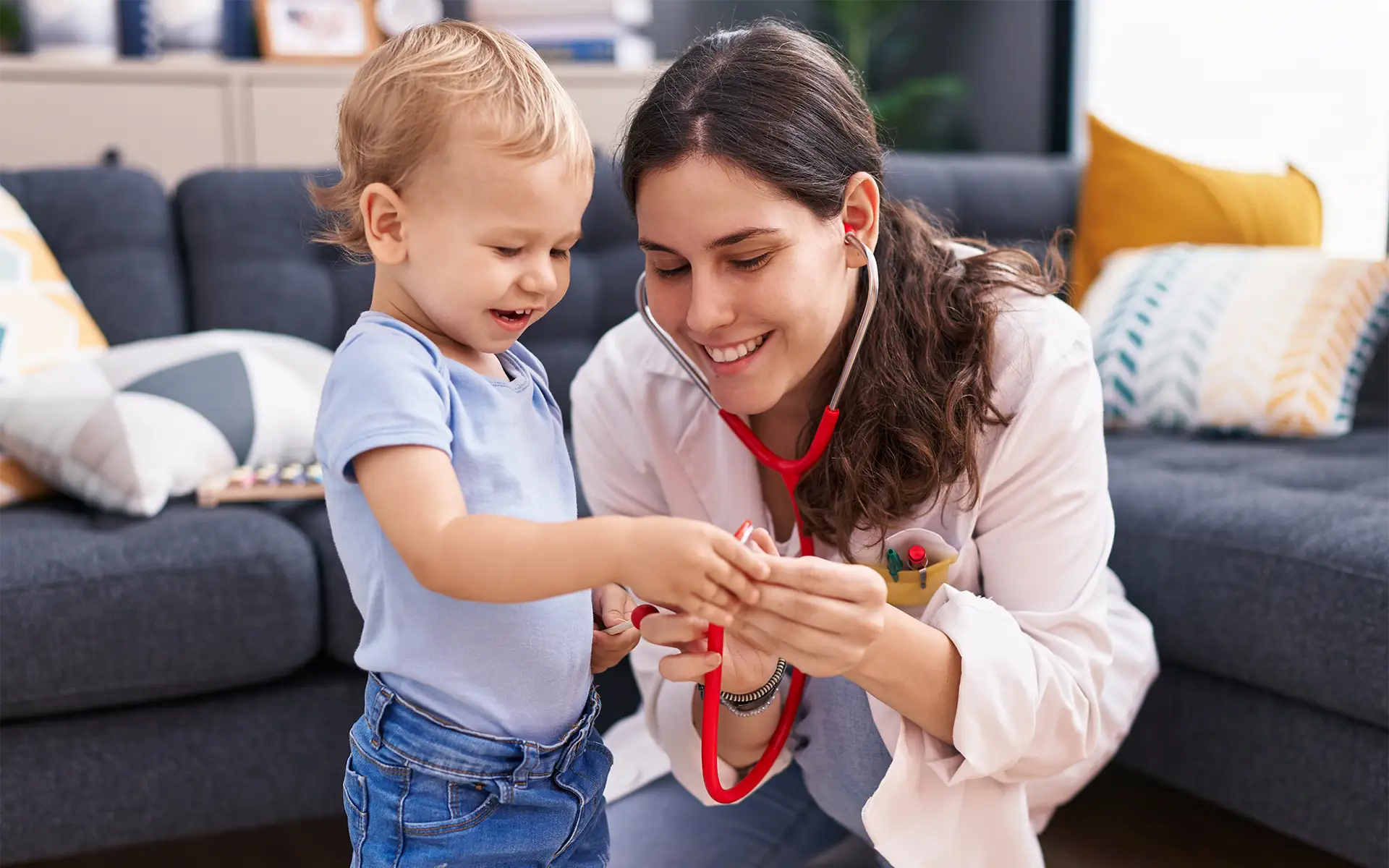 A pediatric nurse kneeling next to a toddler boy in front of a couch, showing him a stethoscope