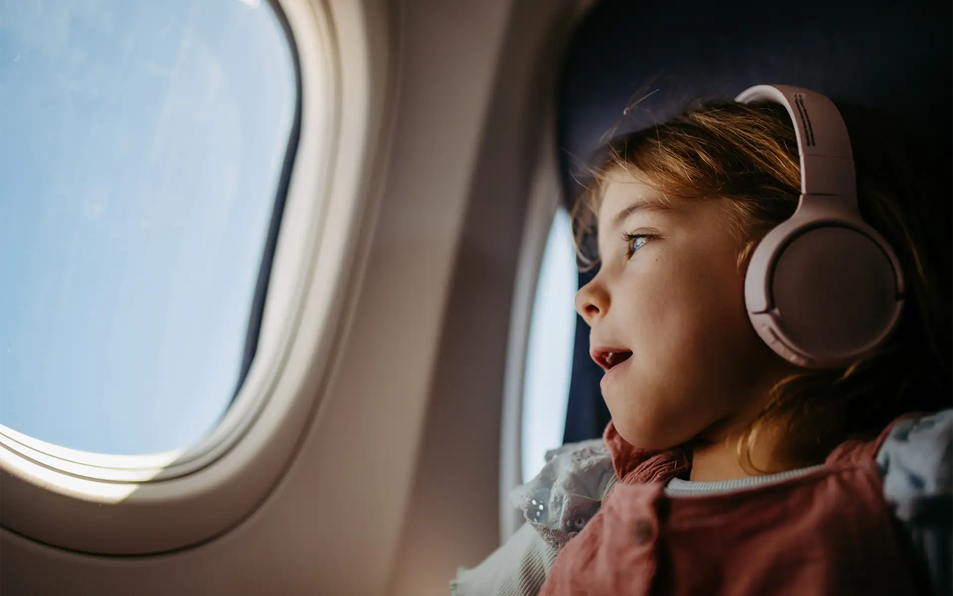 A young girl looks out an airplane window while wearing pink noise-cancelling headphones