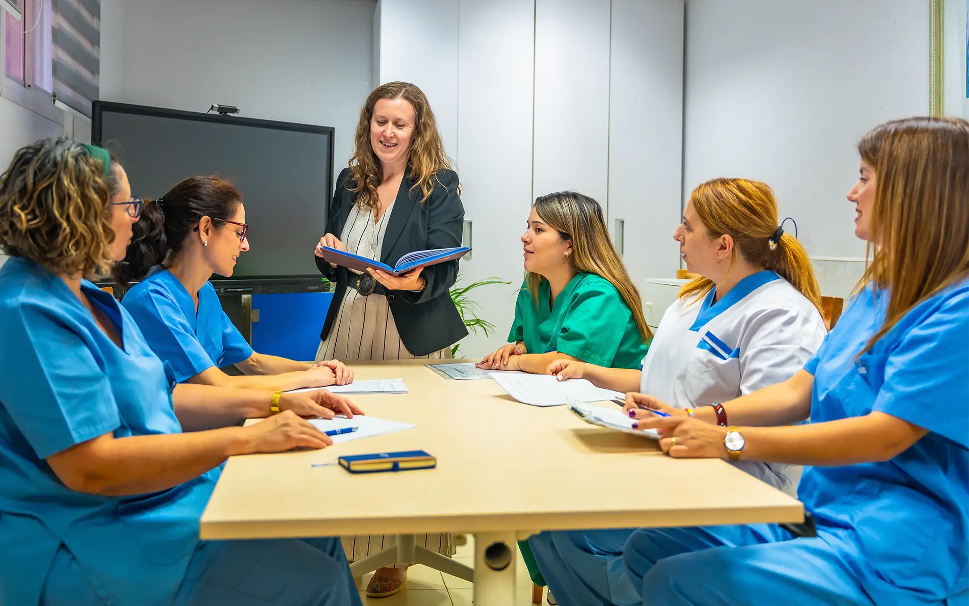 A group of nurses sits around a table, and a female administrator stands during a debriefing to support stress management for nurses