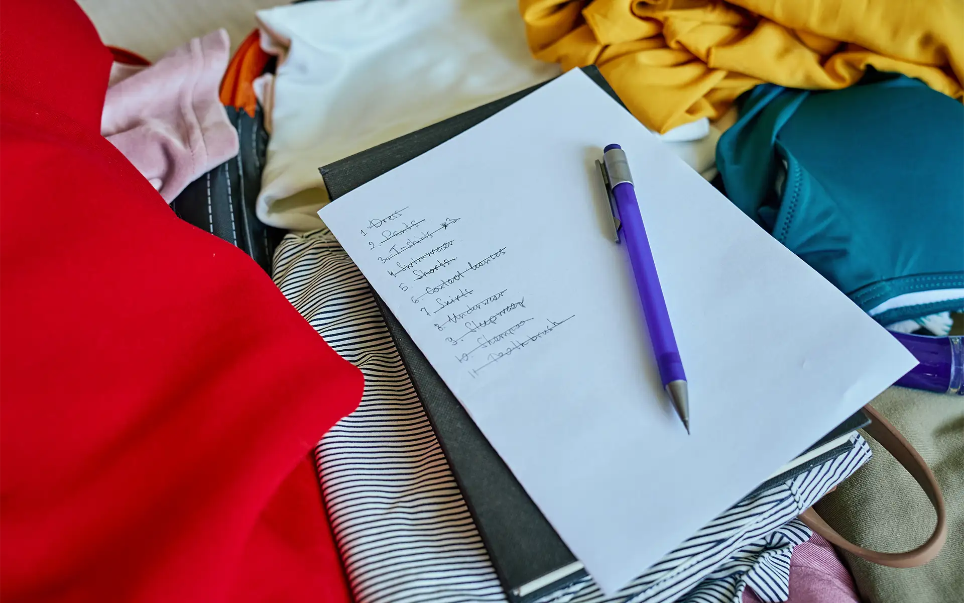 An open suitcase holding clothing with a notepad and pencil resting on top, indicating a packing list