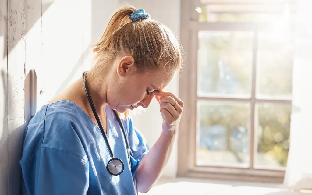 A tired nurse leans against a wall, looking down while pinching the bridge of her nose