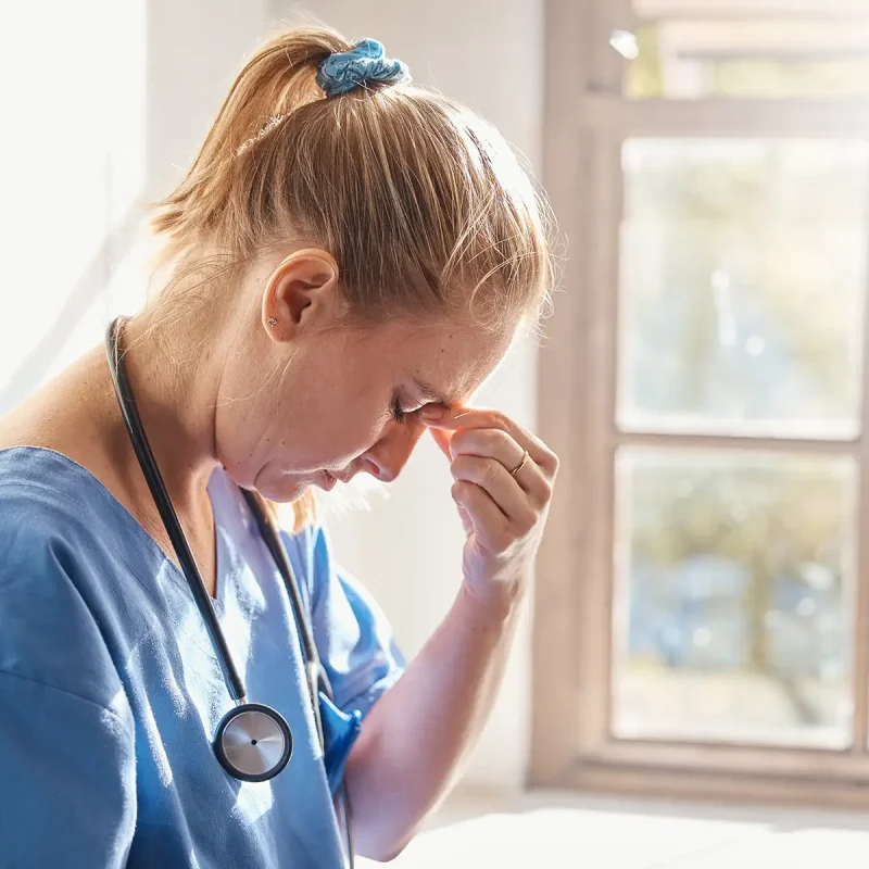 A tired nurse leans against a wall, looking down while pinching the bridge of her nose