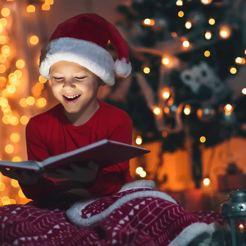 A young boy in a Santa hat sits in front of a decorated holiday tree, reading a book and smiling