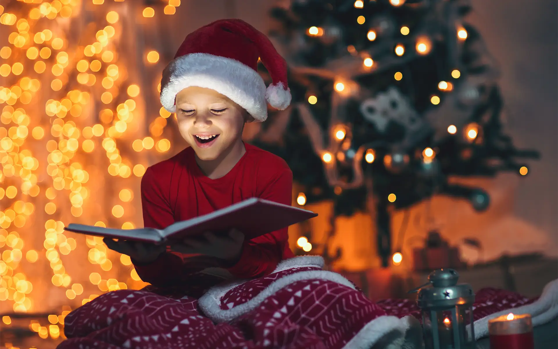 A young boy in a Santa hat sits in front of a decorated holiday tree, reading a book and smiling