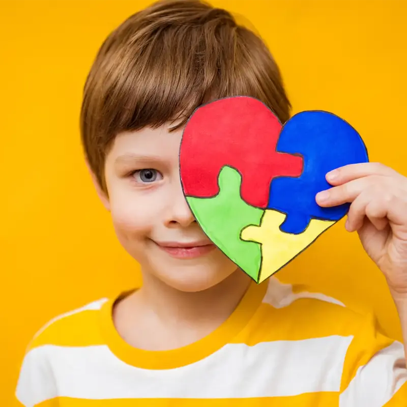 A young boy in a yellow and white striped shirt holds a heart made of colorful puzzle pieces in front of his face
