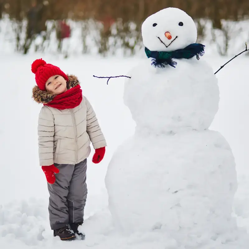Happy young child bundled in winter weather clothing and standing next to a snowman wearing a green scarf