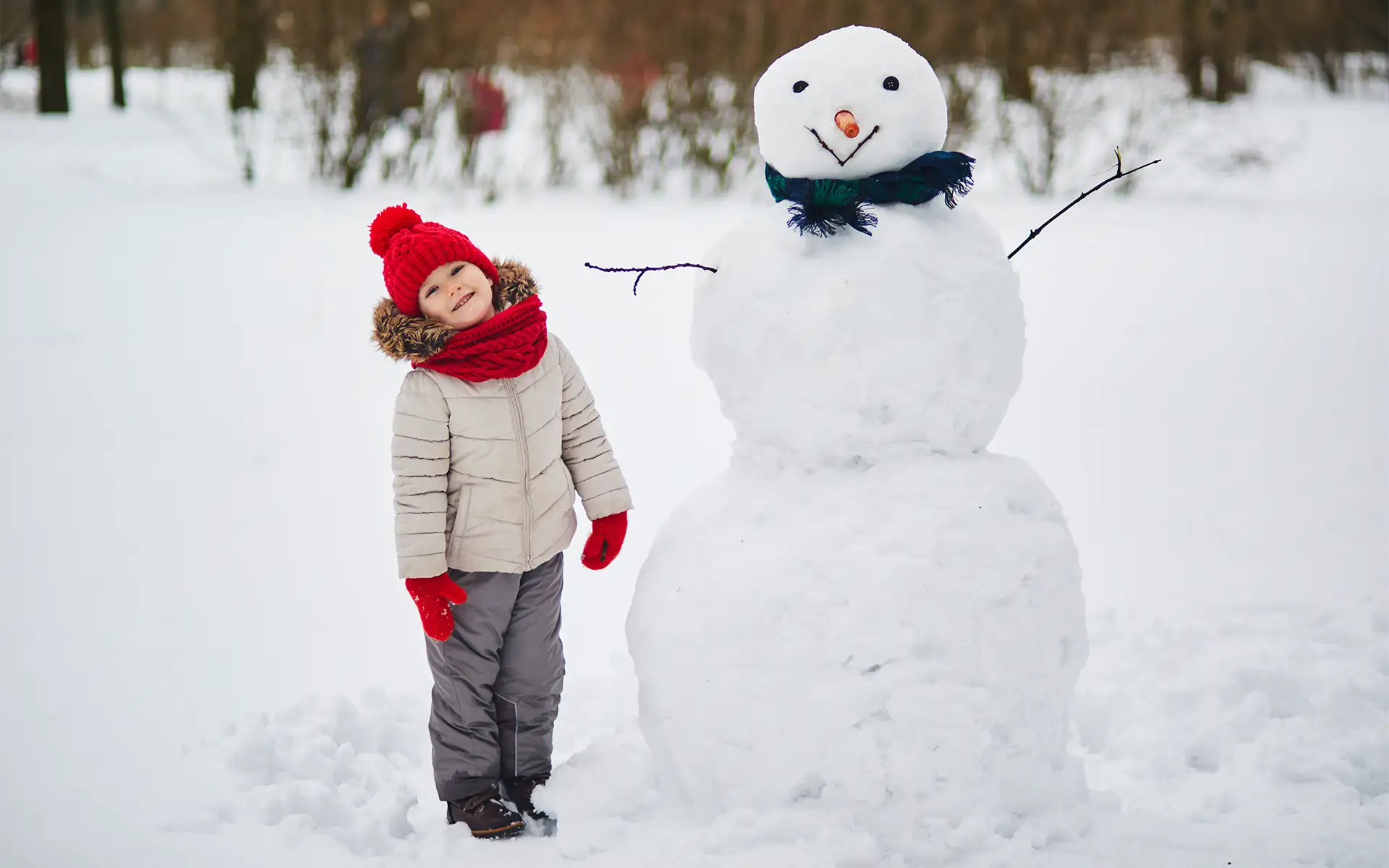Happy young child bundled in winter weather clothing and standing next to a snowman wearing a green scarf