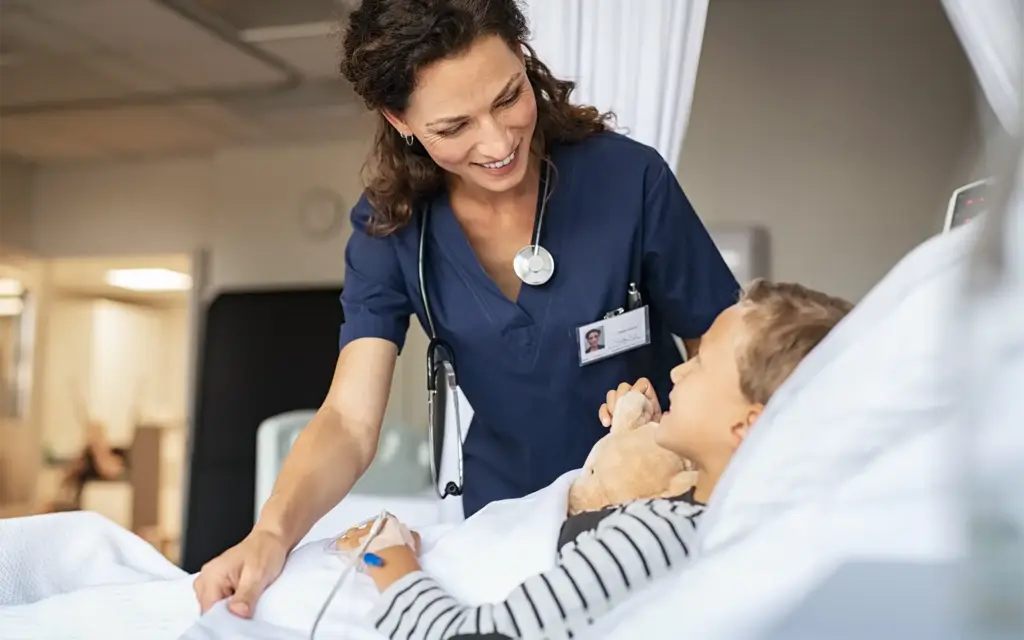 A smiling pediatric nurse stands over a young boy lying in a hospital bed, adjusting his sheet