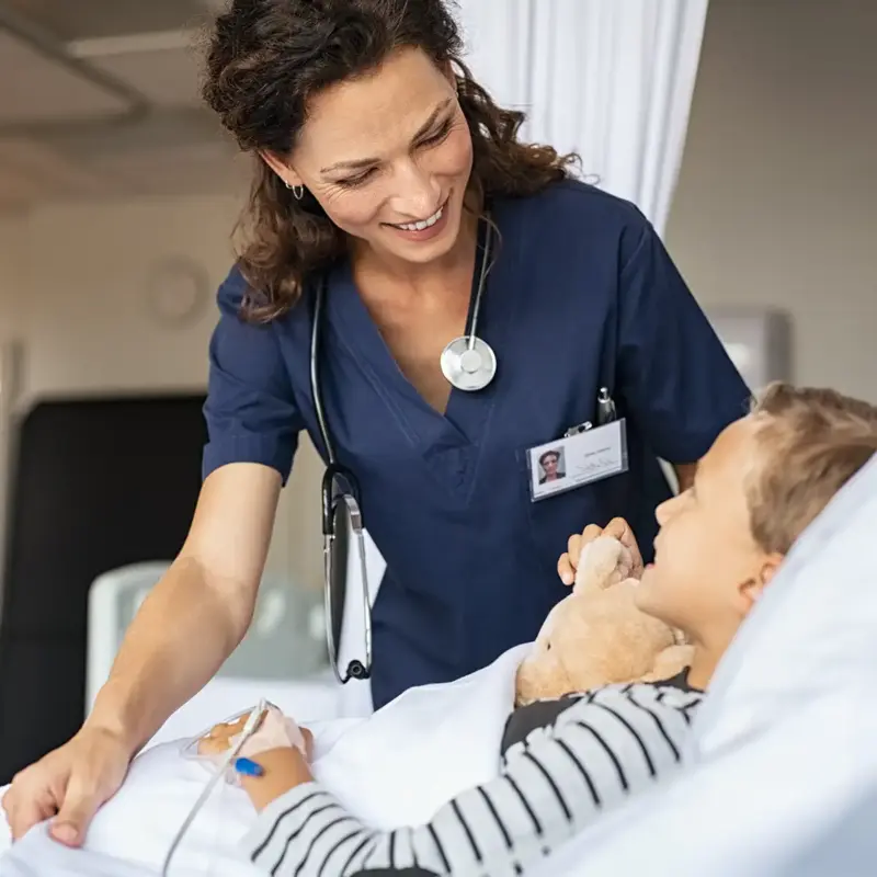A smiling pediatric nurse stands over a young boy lying in a hospital bed, adjusting his sheet