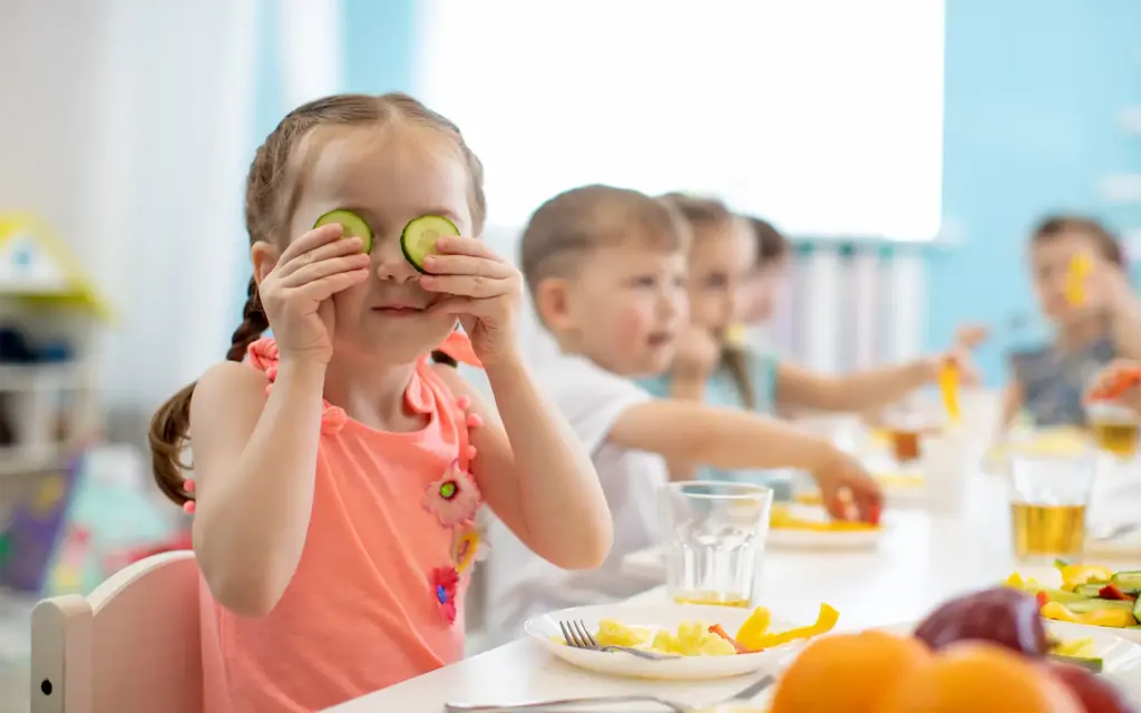A young girl sitting at a lunch table, facing the camera while holding cucumber slices over her eyes