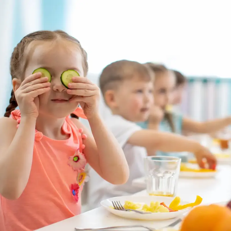 A young girl sitting at a lunch table, facing the camera while holding cucumber slices over her eyes