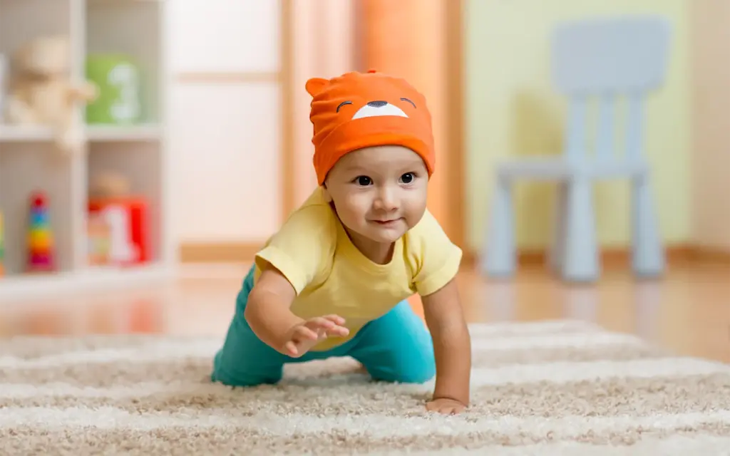 A cute baby in an orange beanie crawling toward the camera on a plush carpet