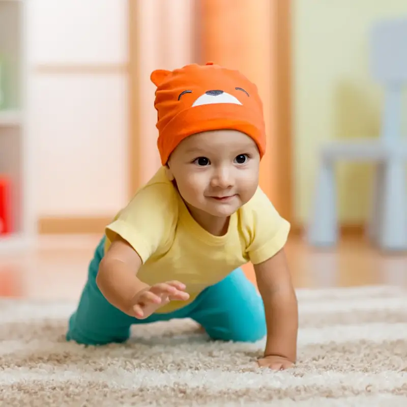 A cute baby in an orange beanie crawling toward the camera on a plush carpet