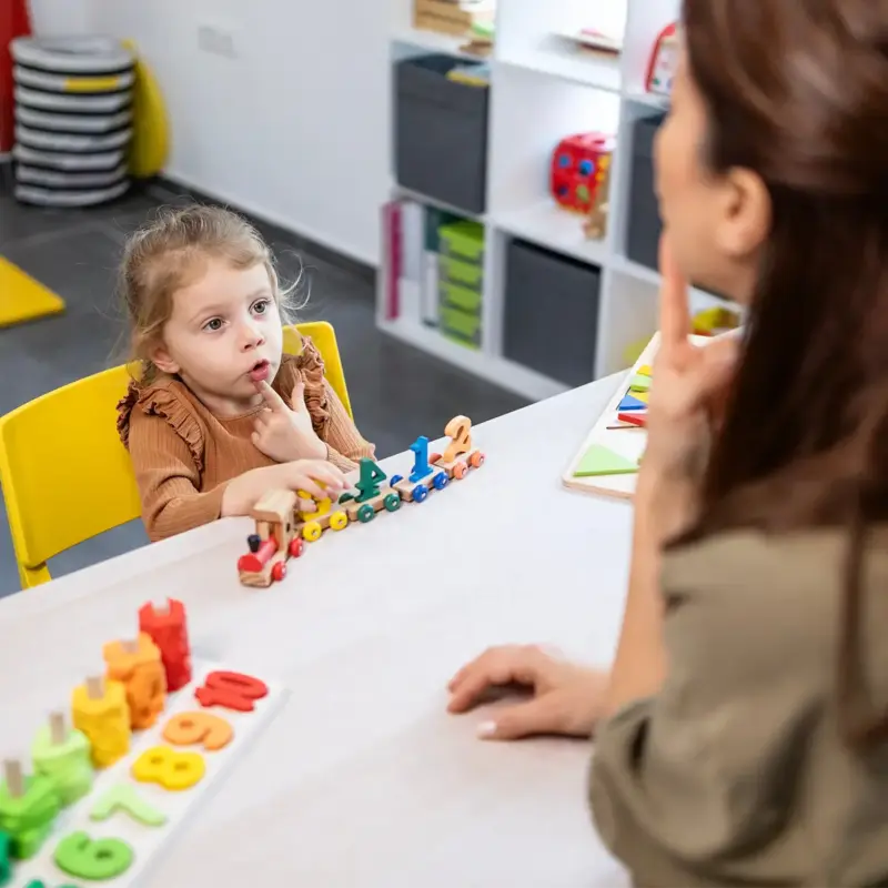 A speech therapist assists a young girl with speech development, engaging in language lessons and communication therapy using educational toys
