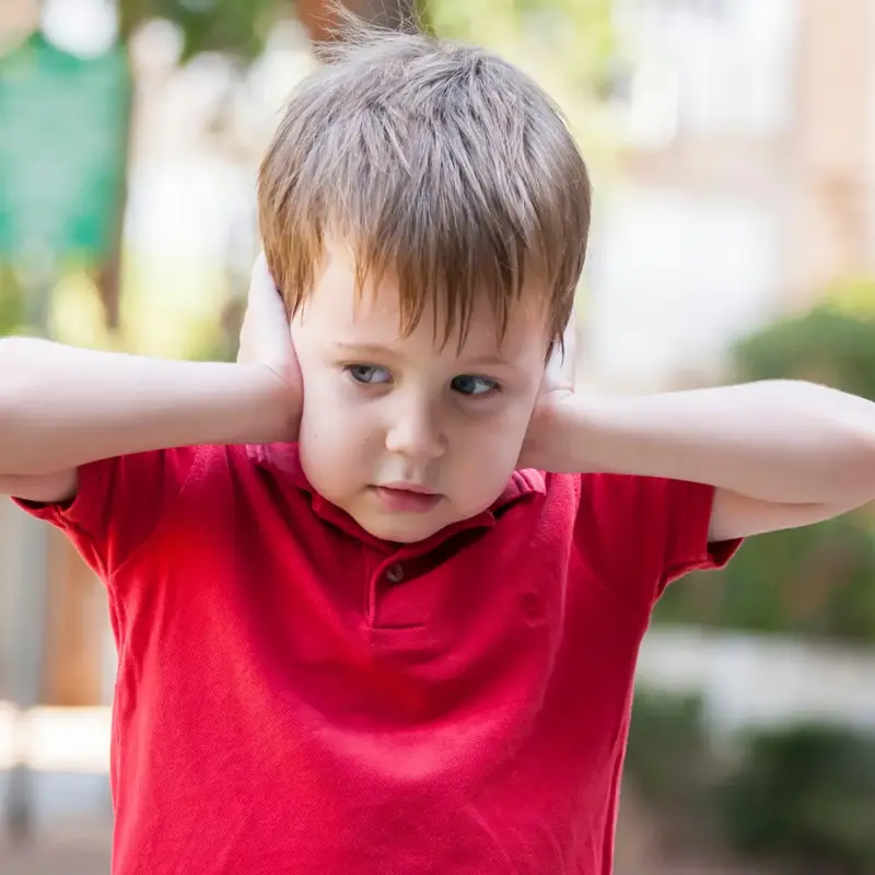 A toddler in a red shirt on a playground covering his ears with his hands