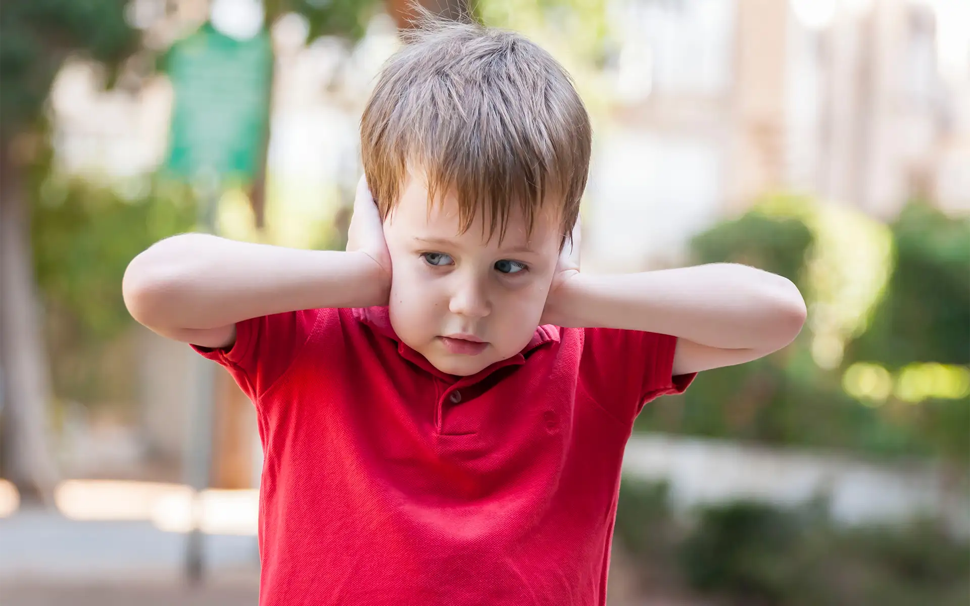 A toddler in a red shirt on a playground covering his ears with his hands