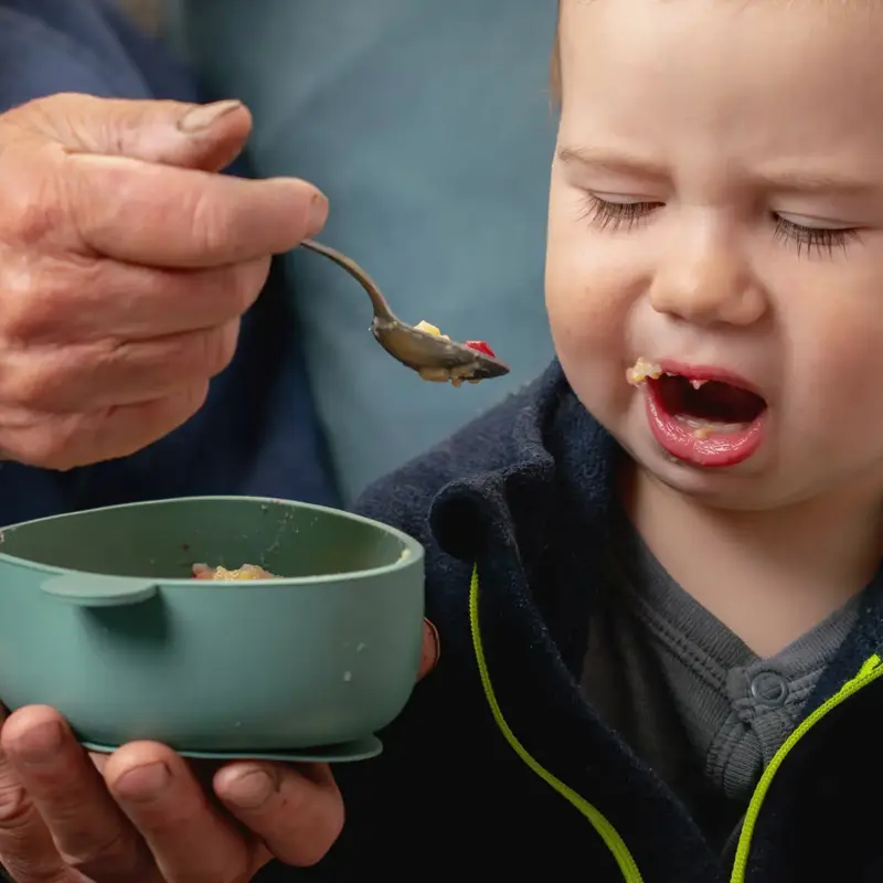 A fussy toddler boy refusing food presented on a spoon by an older caregiver