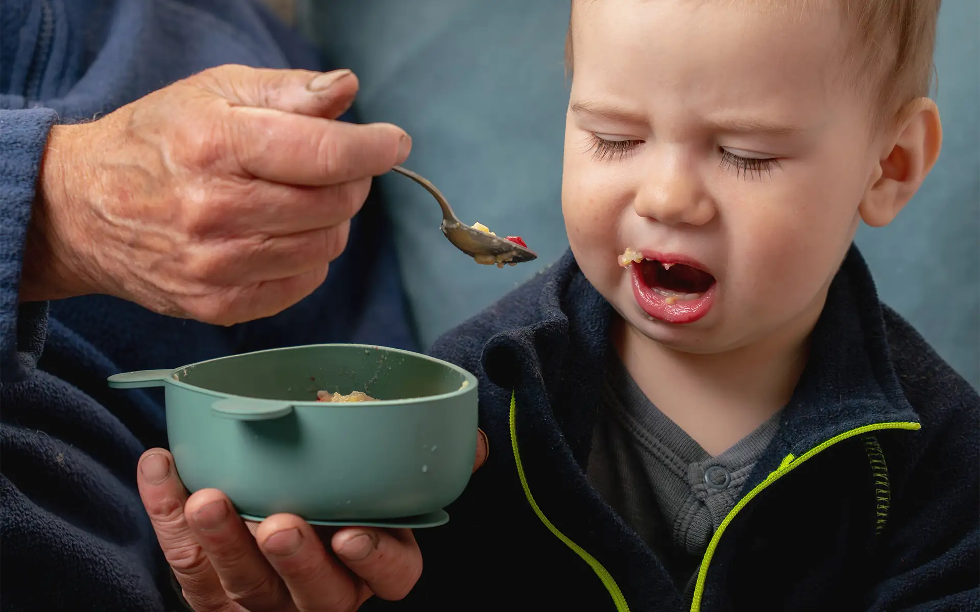 A fussy toddler boy refusing food presented on a spoon by an older caregiver