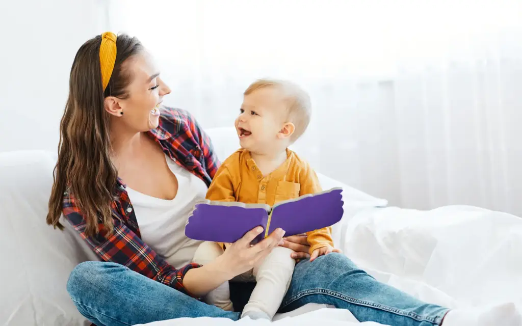 A mother and baby sitting together on a white couch reading a book, looking at each other and smiling
