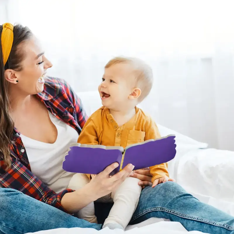 A mother and baby sitting together on a white couch reading a book, looking at each other and smiling