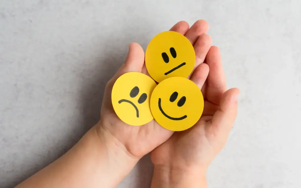 A child's outstretched hands hold three yellow faces featuring a frown, straight face, and smile to signify emotional development in children