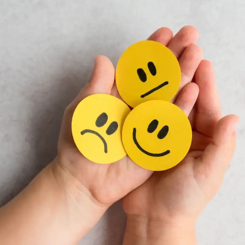 A child's outstretched hands hold three yellow faces featuring a frown, straight face, and smile to signify emotional development in children
