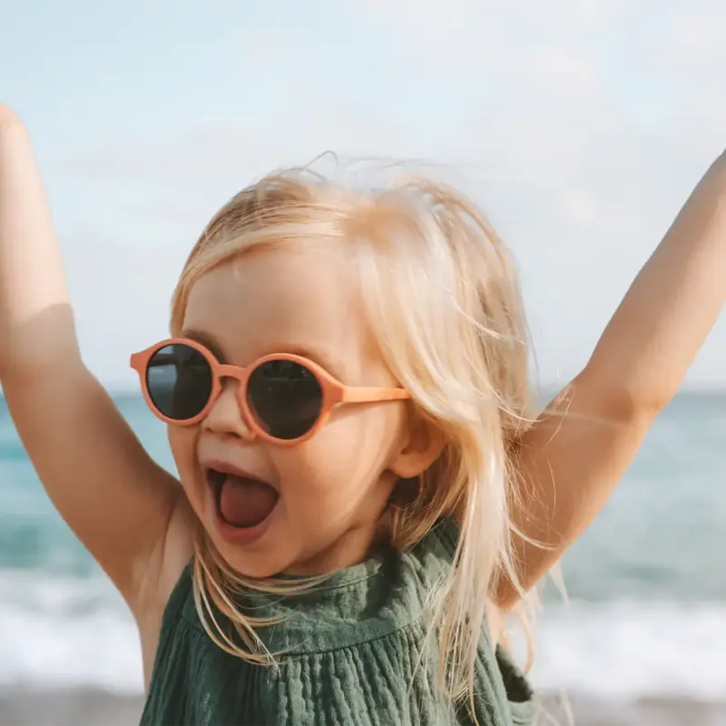 A smiling blonde 3-year-old girl practices UV safety for kids while standing on a beach wearing sunglasses, raising her hands above her head and smiling broadly