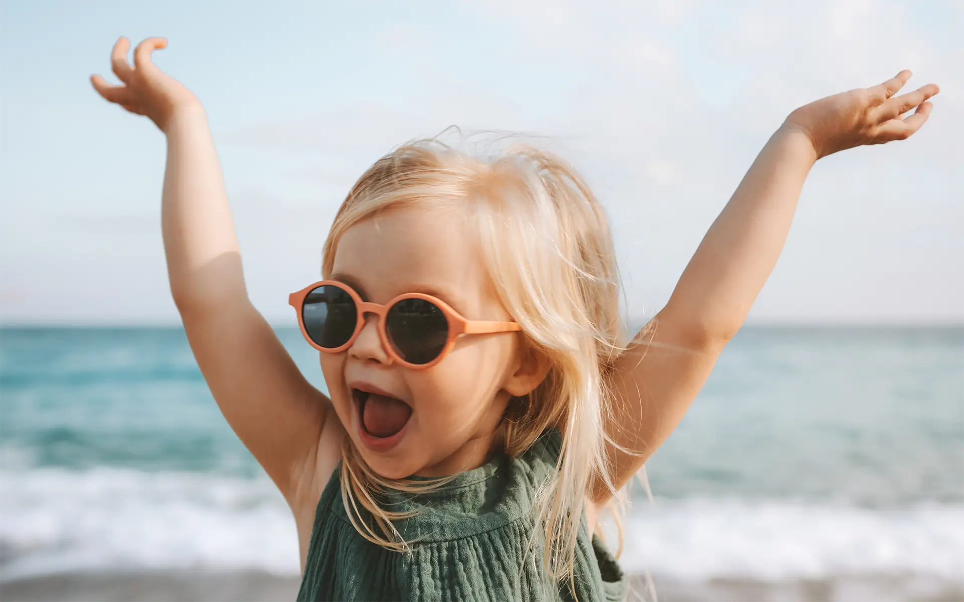 A smiling blonde 3-year-old girl practices UV safety for kids while standing on a beach wearing sunglasses, raising her hands above her head and smiling broadly