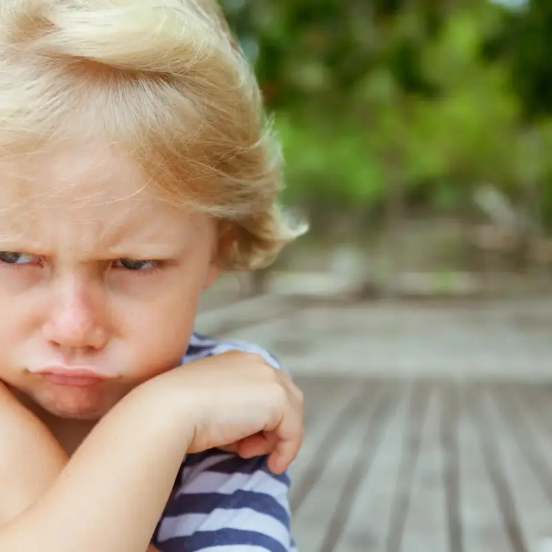 Zoomed-in image of a little blonde boy's frustrated, sad face and crossed arms while standing outside