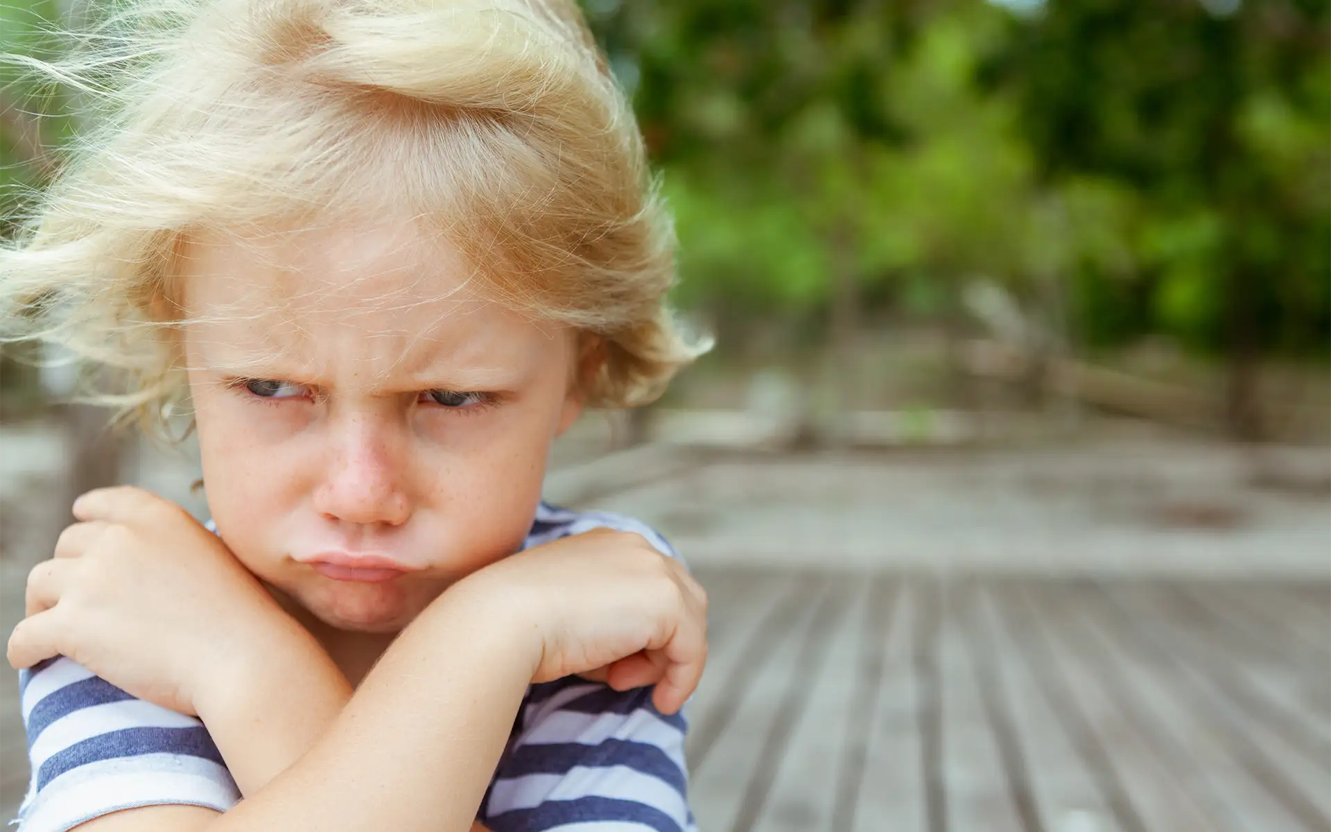 Zoomed-in image of a little blonde boy's frustrated, sad face and crossed arms while standing outside